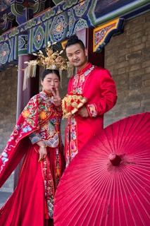 A couple dressed in traditional Chinese wedding attire stand together in front of a decorated structure. The woman is showing a peace sign while the man holds a bouquet. Both are wearing elaborate red and gold outfits with intricate patterns. A large red parasol is placed in front of them, adding to the colorful setting.