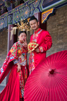 A couple dressed in traditional Chinese wedding attire stand together in front of a decorated structure. The woman is showing a peace sign while the man holds a bouquet. Both are wearing elaborate red and gold outfits with intricate patterns. A large red parasol is placed in front of them, adding to the colorful setting.