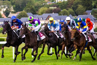 A group of jockeys wearing colorful attire race on horseback across a grassy track. There are multiple horses, each being ridden by a jockey in different vibrant uniforms. In the background, blurred greenery and rooftops of buildings can be seen, indicating a dynamic and fast-paced racing event.