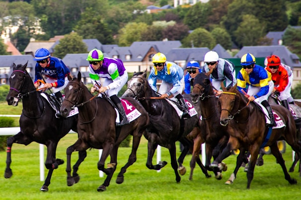 A group of jockeys wearing colorful attire race on horseback across a grassy track. There are multiple horses, each being ridden by a jockey in different vibrant uniforms. In the background, blurred greenery and rooftops of buildings can be seen, indicating a dynamic and fast-paced racing event.