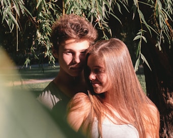 A young couple stands closely together under a tree with hanging branches. The lighting creates soft shadows on their faces, and they appear content and intimate, sharing a quiet moment.