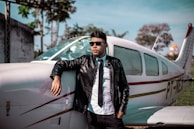 A pilot wearing a check six co. bomber jacket standing confidently beside a small aircraft at sunset.