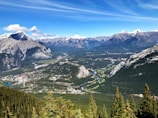 A breathtaking view of a vast mountainous landscape with snow-capped peaks in the distance. A winding river flows through the lush green valley, surrounded by dense forests. A small town with scattered buildings and roads is visible in the lower part of the valley.