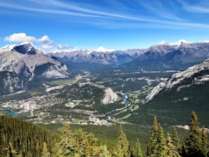A breathtaking view of a vast mountainous landscape with snow-capped peaks in the distance. A winding river flows through the lush green valley, surrounded by dense forests. A small town with scattered buildings and roads is visible in the lower part of the valley.