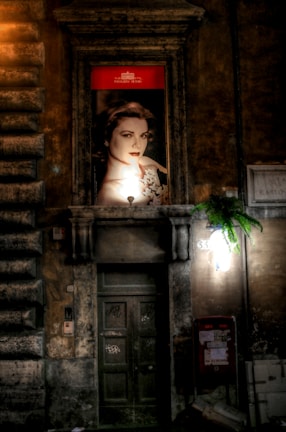 An illuminated advertisement featuring a portrait of a woman is mounted above a rustic, weathered doorway on an old stone building. The background is dimly lit, highlighting the texture of the stone facade. A small potted plant hangs near a light fixture on the right side, adding a touch of greenery.