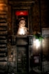 An illuminated advertisement featuring a portrait of a woman is mounted above a rustic, weathered doorway on an old stone building. The background is dimly lit, highlighting the texture of the stone facade. A small potted plant hangs near a light fixture on the right side, adding a touch of greenery.