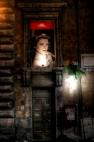 An illuminated advertisement featuring a portrait of a woman is mounted above a rustic, weathered doorway on an old stone building. The background is dimly lit, highlighting the texture of the stone facade. A small potted plant hangs near a light fixture on the right side, adding a touch of greenery.
