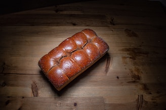 A loaf of freshly baked, golden-brown bread placed on a wooden surface. The bread has a shiny crust with a braided appearance, and it is centered on a rustic table with visible wood grain. Natural lighting casts a shadow to the right.