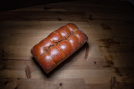 A loaf of freshly baked, golden-brown bread placed on a wooden surface. The bread has a shiny crust with a braided appearance, and it is centered on a rustic table with visible wood grain. Natural lighting casts a shadow to the right.