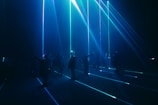 A group of people stand in a dark room with vibrant blue and green laser beams projecting vertically and horizontally around them. The atmosphere is immersive and futuristic, with shadows and light creating a dramatic effect.
