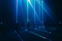 A group of people stand in a dark room with vibrant blue and green laser beams projecting vertically and horizontally around them. The atmosphere is immersive and futuristic, with shadows and light creating a dramatic effect.