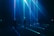 A group of people stand in a dark room with vibrant blue and green laser beams projecting vertically and horizontally around them. The atmosphere is immersive and futuristic, with shadows and light creating a dramatic effect.