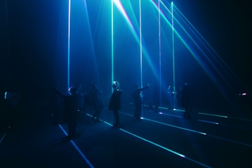A group of people stand in a dark room with vibrant blue and green laser beams projecting vertically and horizontally around them. The atmosphere is immersive and futuristic, with shadows and light creating a dramatic effect.