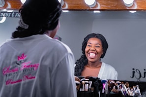 A woman with a joyful expression is looking at herself in a mirror. She is wearing a white robe and has her hair styled neatly. Numerous makeup brushes are arranged in a holder in front of the mirror.