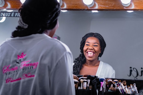 A woman with a joyful expression is looking at herself in a mirror. She is wearing a white robe and has her hair styled neatly. Numerous makeup brushes are arranged in a holder in front of the mirror.