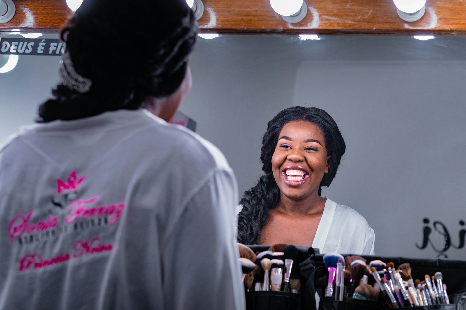 A woman with a joyful expression is looking at herself in a mirror. She is wearing a white robe and has her hair styled neatly. Numerous makeup brushes are arranged in a holder in front of the mirror.