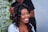 A woman with curly hair is smiling broadly while another person styles her hair with a comb. The background is blurred but appears to have white floral decorations.