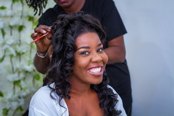 A woman with curly hair is smiling broadly while another person styles her hair with a comb. The background is blurred but appears to have white floral decorations.