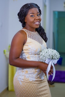 A woman wearing an elegant beige and white dress adorned with lace patterns, smiling while holding a small bouquet of white flowers tied with a satin ribbon. The background features a softly blurred indoor setting.