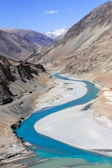 A meandering river with vivid turquoise water flows through a rugged mountainous landscape. The surrounding mountains are arid and barren with shades of brown and grey. Snow-capped peaks are visible in the distance under a clear blue sky, adding to the scenic beauty.