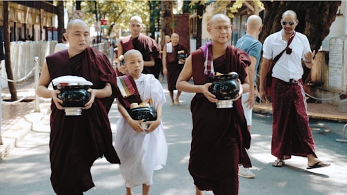 A group of Buddhist monks wearing traditional maroon and white robes walks down a street carrying alms bowls. The background shows other people and trees lining the sidewalk, creating a serene atmosphere.