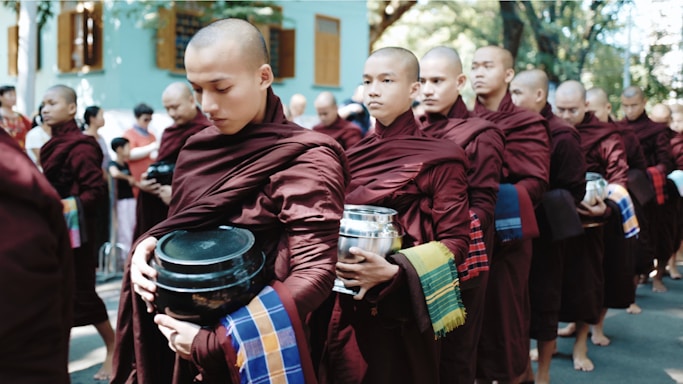 A group of monks dressed in maroon robes holding alms bowls are standing in a single-file line. They appear to be participating in a traditional alms-giving ceremony. The surrounding environment suggests a residential area, with trees and a building in the background.
