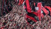 A large crowd of enthusiastic sports fans wave numerous flags in the air. The flags predominantly feature red and black colors, with some having skull motifs. The crowd is densely packed, with many people raising their arms in excitement.