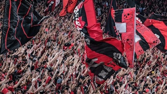 A large crowd of enthusiastic sports fans wave numerous flags in the air. The flags predominantly feature red and black colors, with some having skull motifs. The crowd is densely packed, with many people raising their arms in excitement.