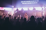 A live music performance with a band on stage, featuring musicians playing instruments like guitar and keyboard. The atmosphere is energetic, as the audience enthusiastically raises their hands, creating a lively and engaging atmosphere. Stage lights illuminate the scene, adding dynamic lighting effects, and lyrics are projected on a screen above the stage.