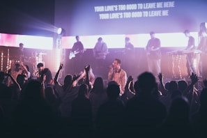 A live music performance with a band on stage, featuring musicians playing instruments like guitar and keyboard. The atmosphere is energetic, as the audience enthusiastically raises their hands, creating a lively and engaging atmosphere. Stage lights illuminate the scene, adding dynamic lighting effects, and lyrics are projected on a screen above the stage.