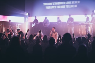 A live music performance with a band on stage, featuring musicians playing instruments like guitar and keyboard. The atmosphere is energetic, as the audience enthusiastically raises their hands, creating a lively and engaging atmosphere. Stage lights illuminate the scene, adding dynamic lighting effects, and lyrics are projected on a screen above the stage.