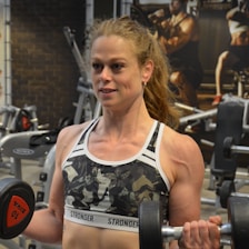 A person is exercising in a gym, performing a bicep curl with a dumbbell in each hand. The individual is wearing a camouflage sports bra that displays the word 'STRONGER'. Various gym equipment is visible in the background, including exercise machines and weight racks.