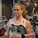 A person is exercising in a gym, performing a bicep curl with a dumbbell in each hand. The individual is wearing a camouflage sports bra that displays the word 'STRONGER'. Various gym equipment is visible in the background, including exercise machines and weight racks.