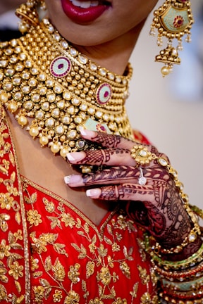 An intricately detailed image featuring a person adorned in elaborate gold jewelry, including a heavily bejeweled necklace and matching earring. The attire is a vibrant red garment with gold floral embroidery. The hand, decorated with intricate henna designs, bares a ring and manicured nails.