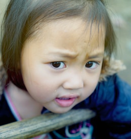 A young child with expressive eyes and furrowed brows, appearing thoughtful or curious. The child is wearing a dark coat with a fur lining, and there is a wooden bar in front of them. The background is blurred, focusing attention on the child's face.
