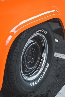 Close-up of a black tire with white lettering on the sidewall, set against an orange fender of a vehicle, with a parking lot surface visible beneath.