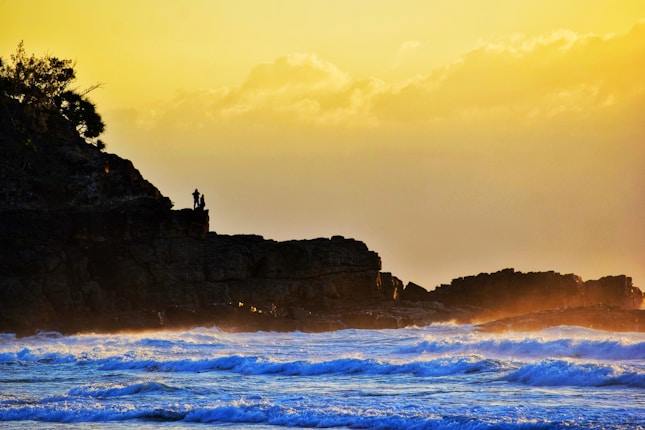A serene coastal scene featuring a rugged cliff with a silhouette of a person standing near the edge. The sky is glowing with a warm golden hue as the sun sets, casting a beautiful light over the ocean waves that crash against the shore.