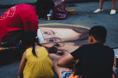 A street artist is creating a large, realistic chalk drawing of a face on the pavement, attracting the attention of two children. The artist wears a red shirt and is focused on the artwork, while the children, one in a yellow dress and the other in dark clothing, watch closely. The surrounding area has a few scattered items, including a small basket and a cup.