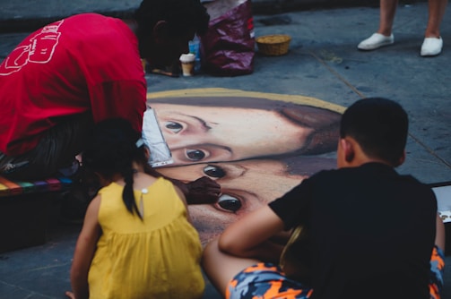 A street artist is creating a large, realistic chalk drawing of a face on the pavement, attracting the attention of two children. The artist wears a red shirt and is focused on the artwork, while the children, one in a yellow dress and the other in dark clothing, watch closely. The surrounding area has a few scattered items, including a small basket and a cup.