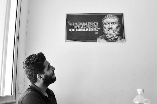 A black and white photograph featuring a man with a beard looking upwards at a framed quote on the wall. The quote is from Plato and is accompanied by a classical-style bust. The setting includes a window on the left and a plastic bottle on a table in the foreground.
