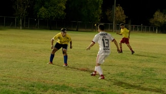 Three soccer players are engaged in an active game on a grassy field at night. One player in a white uniform with 'Tecate' and the number 13 printed on the back is moving towards another player in a yellow jersey and blue shorts, who appears to be in a defensive stance. A third player, also in a yellow jersey and red shorts, is watching the action from a distance. The field is partially lit, and there are trees and a fence in the background.