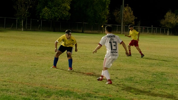 Three soccer players are engaged in an active game on a grassy field at night. One player in a white uniform with 'Tecate' and the number 13 printed on the back is moving towards another player in a yellow jersey and blue shorts, who appears to be in a defensive stance. A third player, also in a yellow jersey and red shorts, is watching the action from a distance. The field is partially lit, and there are trees and a fence in the background.