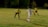 Three soccer players are engaged in an active game on a grassy field at night. One player in a white uniform with 'Tecate' and the number 13 printed on the back is moving towards another player in a yellow jersey and blue shorts, who appears to be in a defensive stance. A third player, also in a yellow jersey and red shorts, is watching the action from a distance. The field is partially lit, and there are trees and a fence in the background.