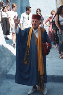 An elderly man wearing traditional clothing, including a red fez and a blue robe with a yellow scarf, waves while standing on a street. Several people are in the background, some looking towards him, others engaged in their own activities. The buildings and surroundings are painted in varying shades of blue, suggesting a location known for such architecture.