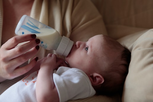 A baby is lying down while being fed with a milk bottle held by an adult hand. The infant is wearing a white shirt and is resting on a beige cushion. The adult's nails are painted a dark red color.