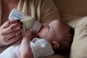A baby is lying down while being fed with a milk bottle held by an adult hand. The infant is wearing a white shirt and is resting on a beige cushion. The adult's nails are painted a dark red color.