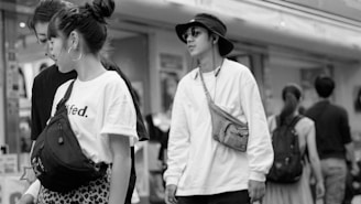 A black and white urban scene capturing several people walking leisurely. The primary focus is on a young woman with braided hair wearing a graphic t-shirt and a sling bag, and a young man in sunglasses with a casual hat and a crossbody bag. They seem to be conversing or interacting with each other. The background shows a busy street with more people walking, and a storefront is visible.