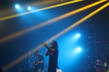 A live concert scene with two musicians performing on stage. One is holding a microphone and engaging with the audience, while the other plays a guitar. Bright stage lights in blue and yellow cast beams, creating a dynamic and energetic atmosphere.