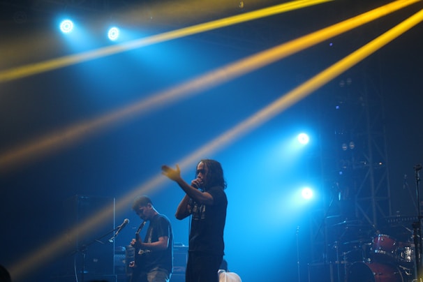 A live concert scene with two musicians performing on stage. One is holding a microphone and engaging with the audience, while the other plays a guitar. Bright stage lights in blue and yellow cast beams, creating a dynamic and energetic atmosphere.