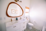 A modern bathroom with a large wooden-framed mirror above a dual sink vanity. The sinks have gold faucets, and the vanity is white with golden handles. A toilet is situated next to the sink, beneath a window covered with a woven blind. The floor features a green and white geometric pattern. Two framed pictures hang on the wall.